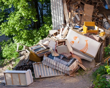 The image shows a large pile of mixed household rubbish and debris situated outdoors, likely in a yard or alleyway near a residential area. The waste includes various wooden furniture pieces such as a striped upholstered armchair, a small wooden table, and a white chest of drawers with visible handles, all stacked in a disorderly manner. There are also cardboard boxes, some with printed or painted designs, and other miscellaneous waste materials like pieces of wood and dismantled furniture parts. The debris appears weathered, with some items exhibiting signs of exposure to the elements, and the pile is positioned against a background of greenery with leafy trees, suggesting an outdoor environment in Tooting or a nearby postcode. A large, corrugated metal sheet leans against the wall or fence behind the heap, indicating an industrial or storage area. The scene is illuminated by natural daylight, highlighting the textures and variety of materials within the rubbish. This image illustrates a typical scenario where Commercial Waste Tooting would assist with professional rubbish removal and clearance services in the local area.