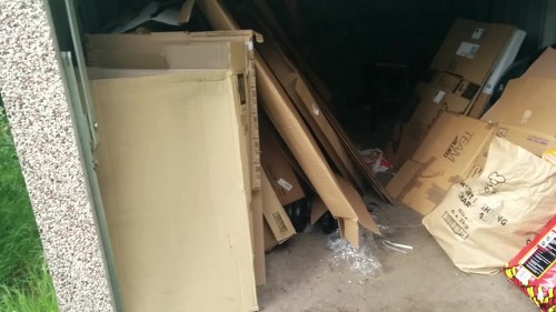 A cluttered indoor or garage space with a variety of discarded items including a large cream-colored wooden wardrobe or cabinet leaning to the side, alongside numerous cardboard boxes and packaging materials, some with printed labels or logos, scattered and stacked haphazardly on the floor. The surface appears to be concrete or similar material, with some plastic wrapping and packaging debris visible. The area is dimly lit and shows signs of recent clearing or rubbish collection, reflecting typical rubbish removal activities that a professional service such as Commercial Waste Tooting might handle for local businesses or residents. Background elements include darker interior space and additional boxes, with a glimpse of a domestic or commercial environment suitable for waste disposal and clearance services in Tooting or nearby postcode districts.