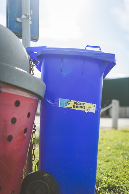 A row of large, rectangular wheelie bins lined up along a paved sidewalk outdoors, positioned next to a brick wall. The bins are made of durable plastic with textured surfaces and are painted in vario